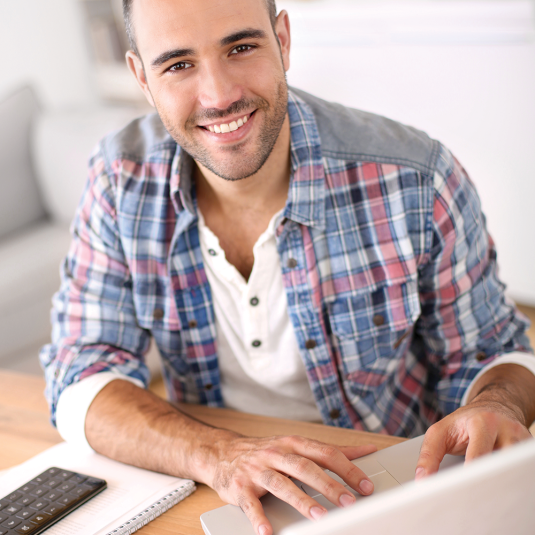 A smiling man sits at a table, focused and content, engaged with his laptop.