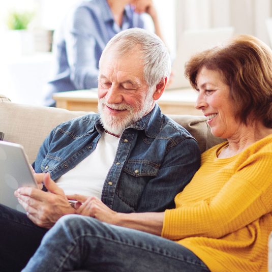 An older couple sits on the couch, sharing a moment of connection while using a tablet together.